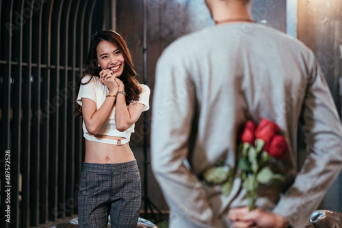 Man hiding roses flower to surprise his girlfriend on dating in Valentine's, Anniversary or birthday, focus on stanging happy woman smile on face, attractive couple having romantic on rooftop photo