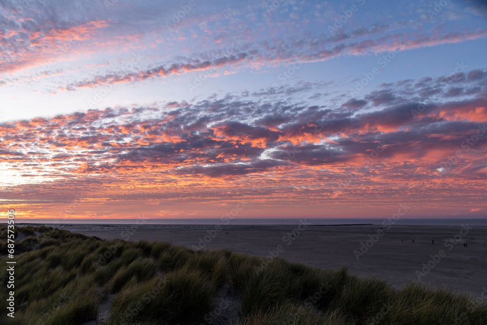 Panoramic sunset view over the dunes on the island of Texel in the Netherlands with long bands of orange coloring clouds in sky with dune grass in forefront and people walking on the wide beach Panoramic sunset view over the dunes on the island of Texel in the Netherlands with long bands of orange coloring clouds in sky with dune grass in forefront and people walking on the wide beach