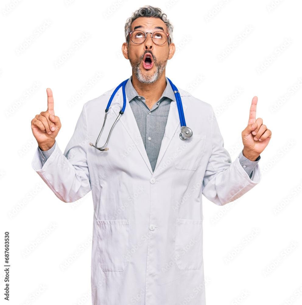 Middle age grey-haired man wearing doctor uniform and stethoscope amazed and surprised looking up and pointing with fingers and raised arms. Middle age grey-haired man wearing doctor uniform and stethoscope amazed and surprised looking up and pointing with fingers and raised arms.