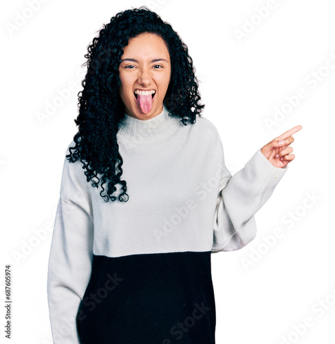 Young hispanic woman with curly hair pointing with fingers to the side sticking tongue out happy with funny expression. photo