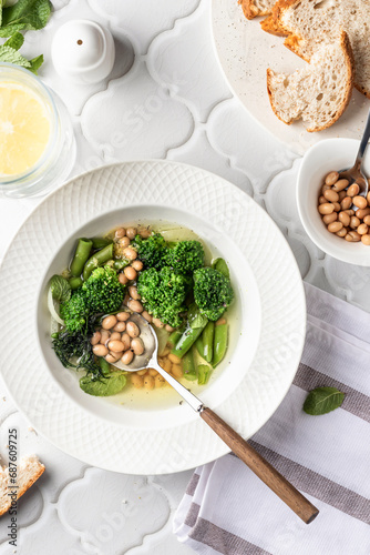 Vegetarian minestrone soup with green vegetables and beans served in white plate on white tile background, top view