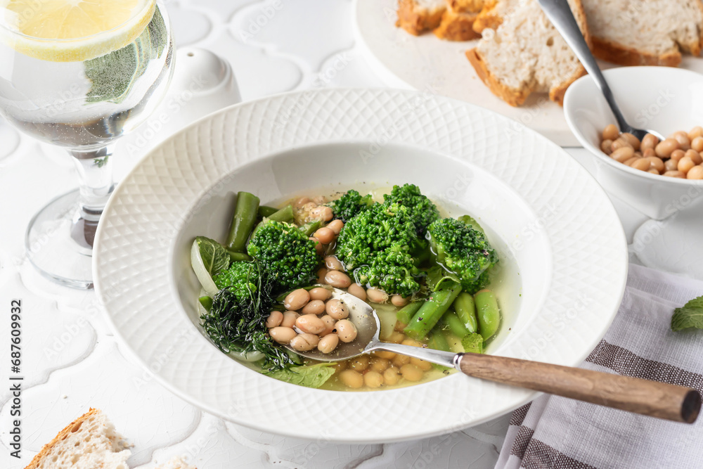 Vegetarian minestrone soup with green vegetables and beans served in white plate and bread on white tile background