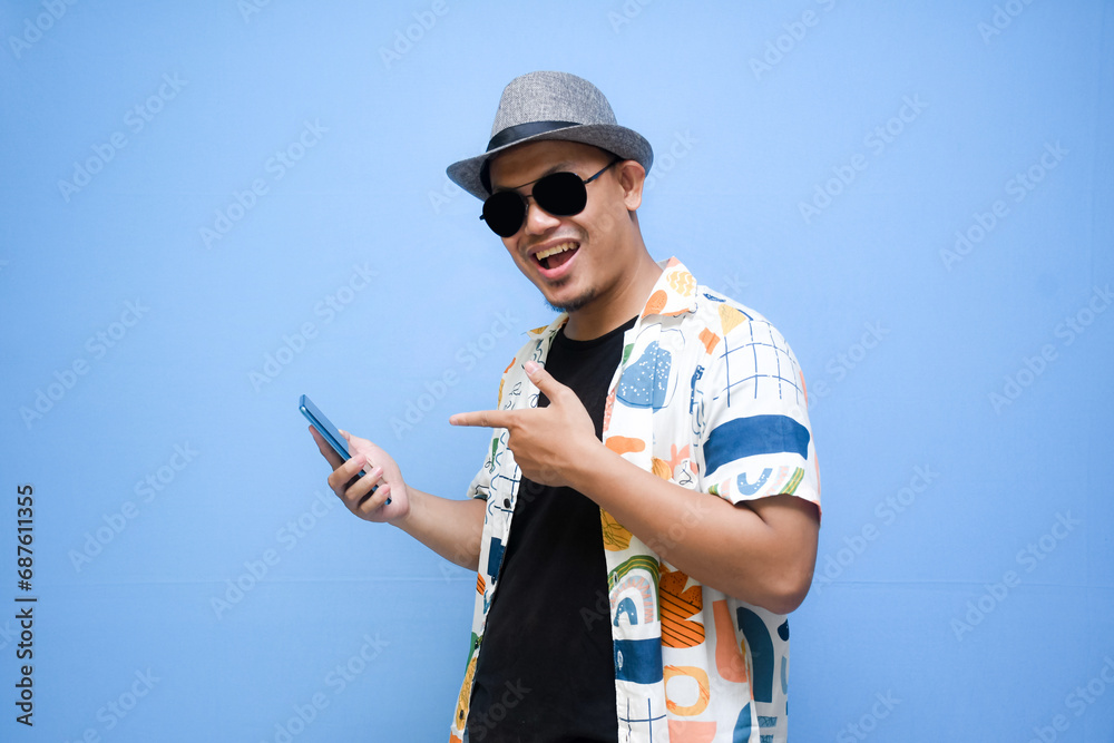 Happy asian man wearing beach shirt holding a phone while pointing a finger standing over blue background. Vacation concept