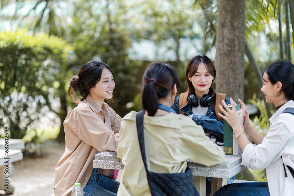 Group of young Asian college students sitting on a bench in a campus relaxation area, talking, sharing ideas, doing homework or tutoring for the exam together Group of young Asian college students sitting on a bench in a campus relaxation area, talking, sharing ideas, doing homework or tutoring for the exam together