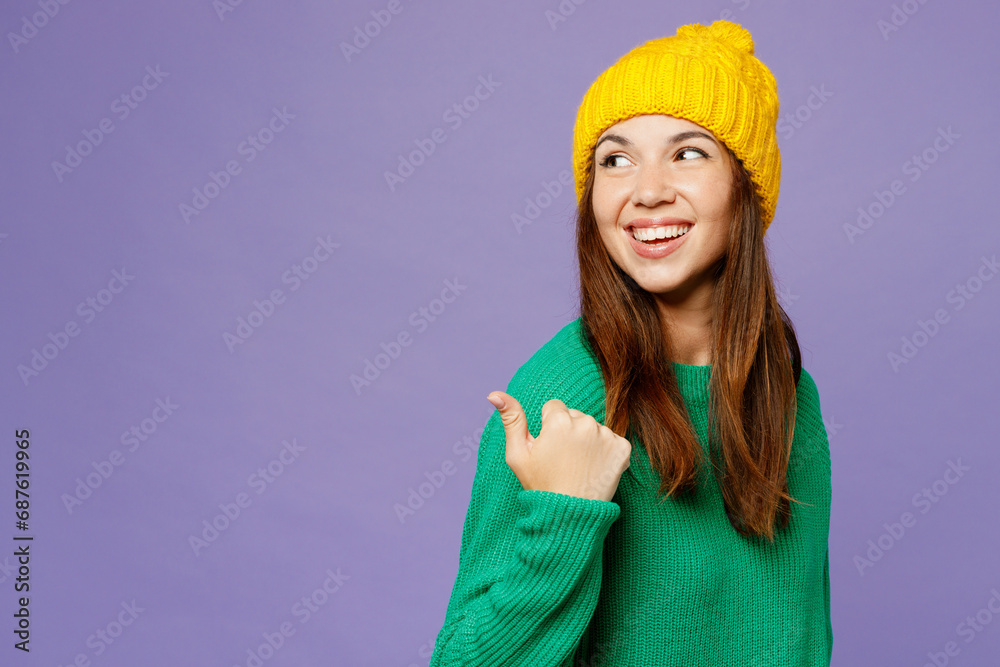 Side view young woman she wearing green sweater yellow hat casual clothes point thumb finger aside on area mock up isolated on plain pastel light purple background studio portrait. Lifestyle concept.