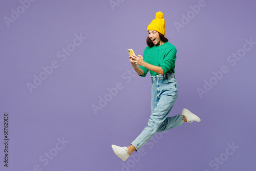 Full body young woman she wear green sweater yellow hat casual clothes jump high hold in hand use mobile cell phone run fast isolated on plain pastel light purple background studio. Lifestyle concept.