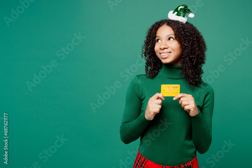 Merry fun little kid teen girl wear hat casual clothes posing hold credit bank card look aside isolated on plain green background studio portrait. Happy New Year celebration Christmas holiday concept.