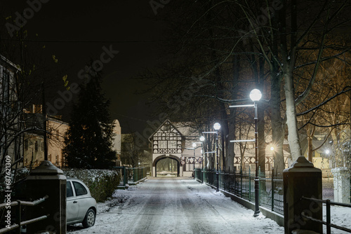 Ulica Pałacowa w miejscowości Iłowa w Polsce. Jest zimowa noc, jezdnię i chodnik pokrywa warstwa śniegu, ciemności rozświetlają latarnie. Ulica zakończona jest bramą w piętrowym budynku.