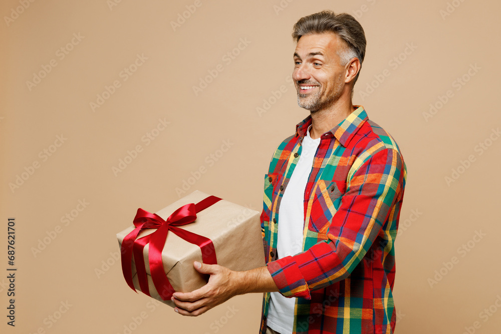 Adult happy man wear red shirt white t-shirt casual clothes hold in hand give present box with gift ribbon bow isolated on plain pastel light beige color background studio portrait. Lifestyle concept.