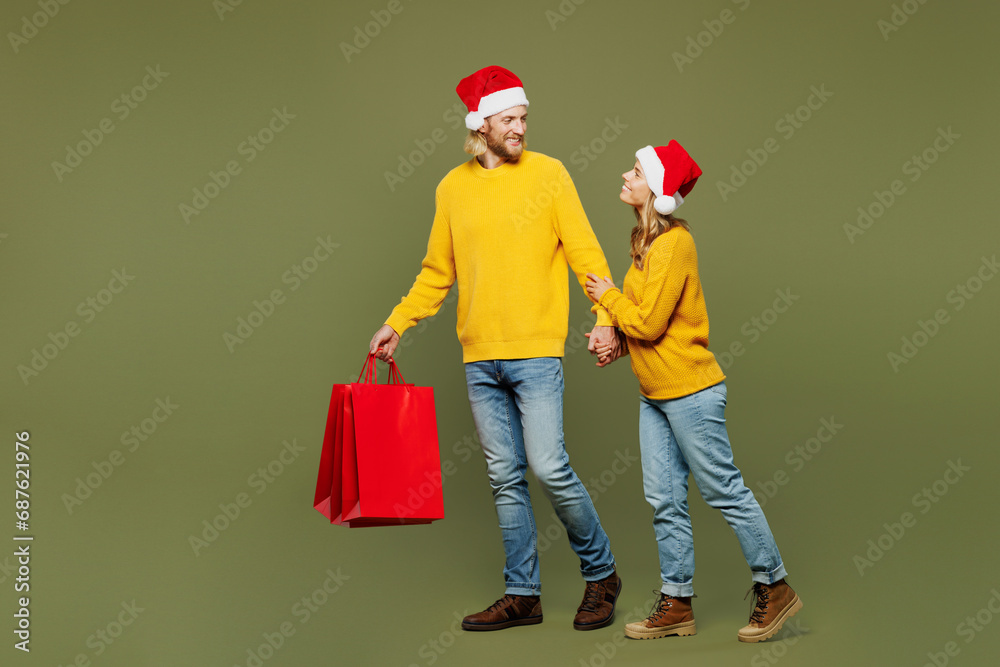 Fototapeta premium Full body sideways merry young couple two friends man woman wears sweater Santa hat posing hold shopping bags isolated on plain light green background. Happy New Year Christmas sale buy day concept.