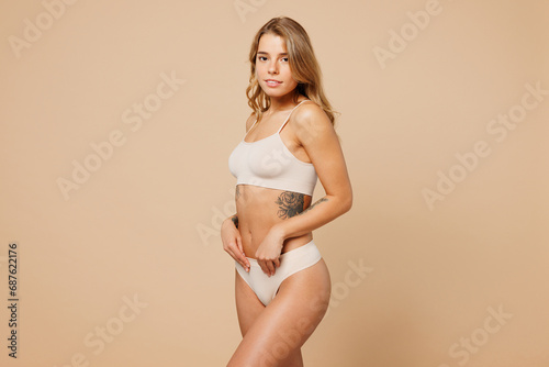 Side view young nice lady woman with slim body perfect skin wear nude top bra lingerie stand put hand on belly look camera isolated on plain pastel light beige background. Lifestyle diet fit concept