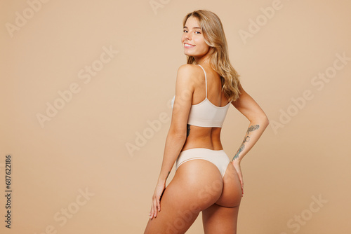 Back side view young nice lady woman with slim body perfect skin wear nude top bra lingerie stand look camera put hand on thigh isolated on plain pastel beige background. Lifestyle diet fit concept.