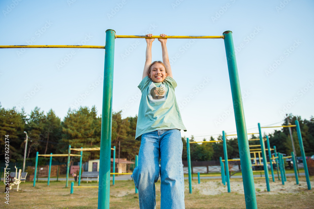 Fototapeta premium Girl hanging on horizontal bar. Child doing motion exercises and playing on playground outdoor. Healthy leisure sport activity on fresh air. Happy childhood. Kids physical training on sports stadium