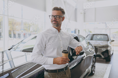 Adult man customer buyer client wear shirt hold takeaway delivery cup coffee to go loot at watch choose auto wants buy new automobile in car showroom salon dealership store motor show. Sales concept.