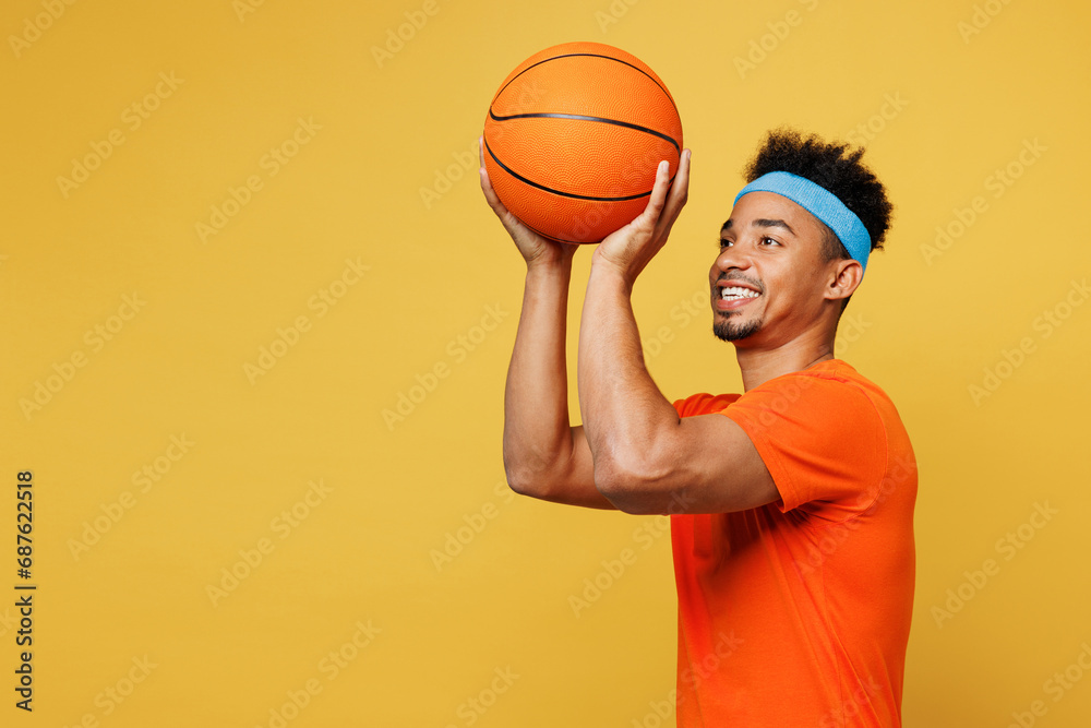 Naklejka premium Young fitness trainer instructor sporty man sportsman wear orange t-shirt hold in hand basketball ball shooting training in home gym isolated on plain yellow background. Workout sport fit abs concept.