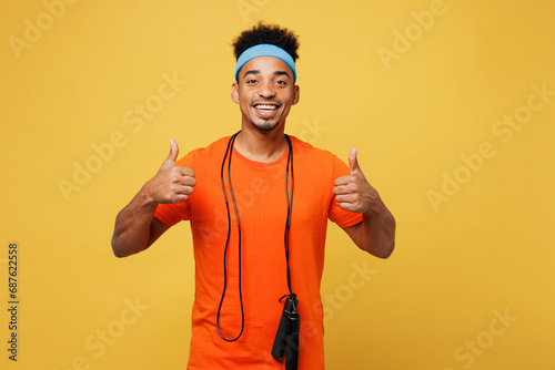 Young fitness trainer instructor sporty man sportsman wears orange t-shirt hold skipping rope show thumb up spend time in home gym isolated on plain yellow background. Workout sport fit abs concept. photo