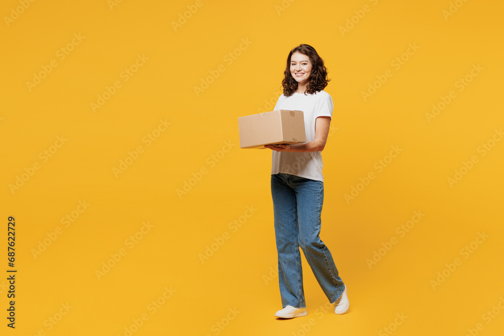 Fototapeta premium Full body side view young happy courier woman she wears white blank t-shirt casual clothes hold cardboard box look camera isolated on plain yellow orange background studio portrait. Lifestyle concept.