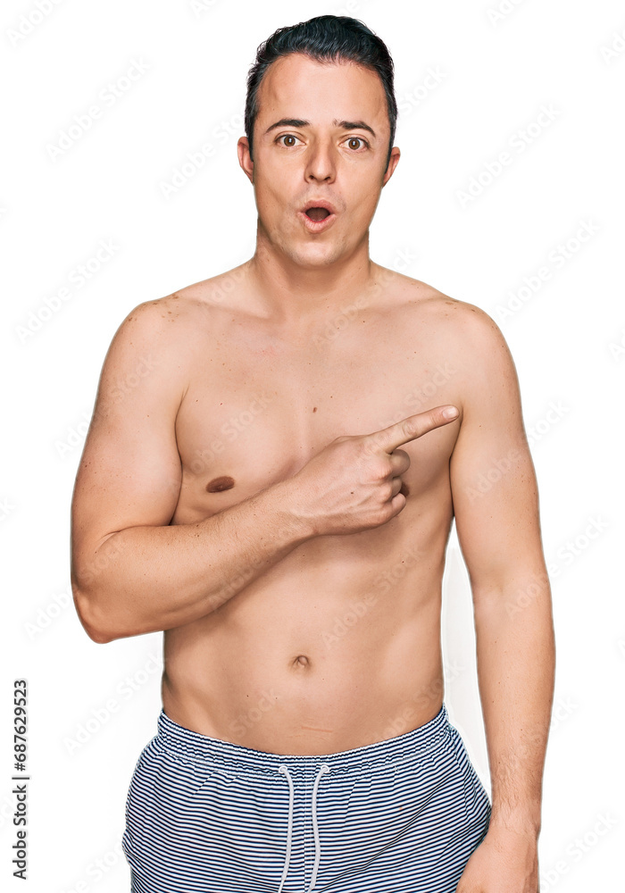 Fototapeta premium Handsome young man wearing swimwear shirtless surprised pointing with finger to the side, open mouth amazed expression.