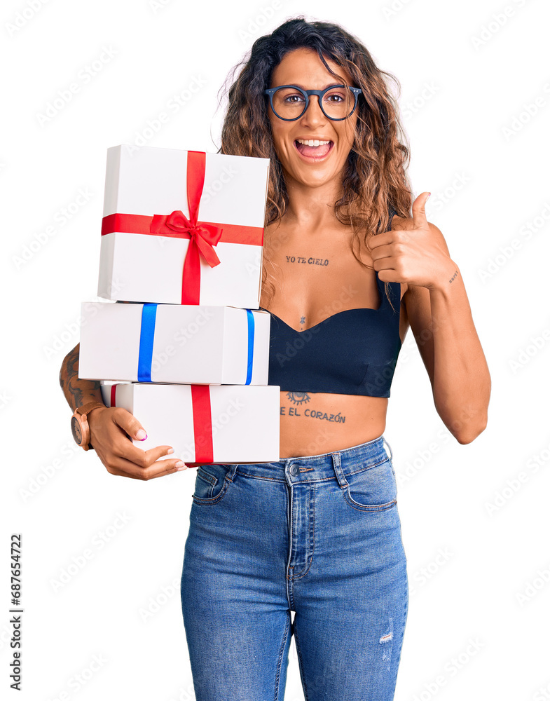 Young hispanic woman with tattoo holding gifts smiling happy and positive, thumb up doing excellent and approval sign Young hispanic woman with tattoo holding gifts smiling happy and positive, thumb up doing excellent and approval sign