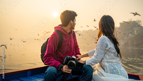 Young couple looking at the sunrise or sunset while sitting on the boat, holding a camera in hand, Asian partners enjoying their vacation together, Indian lovers on a trip