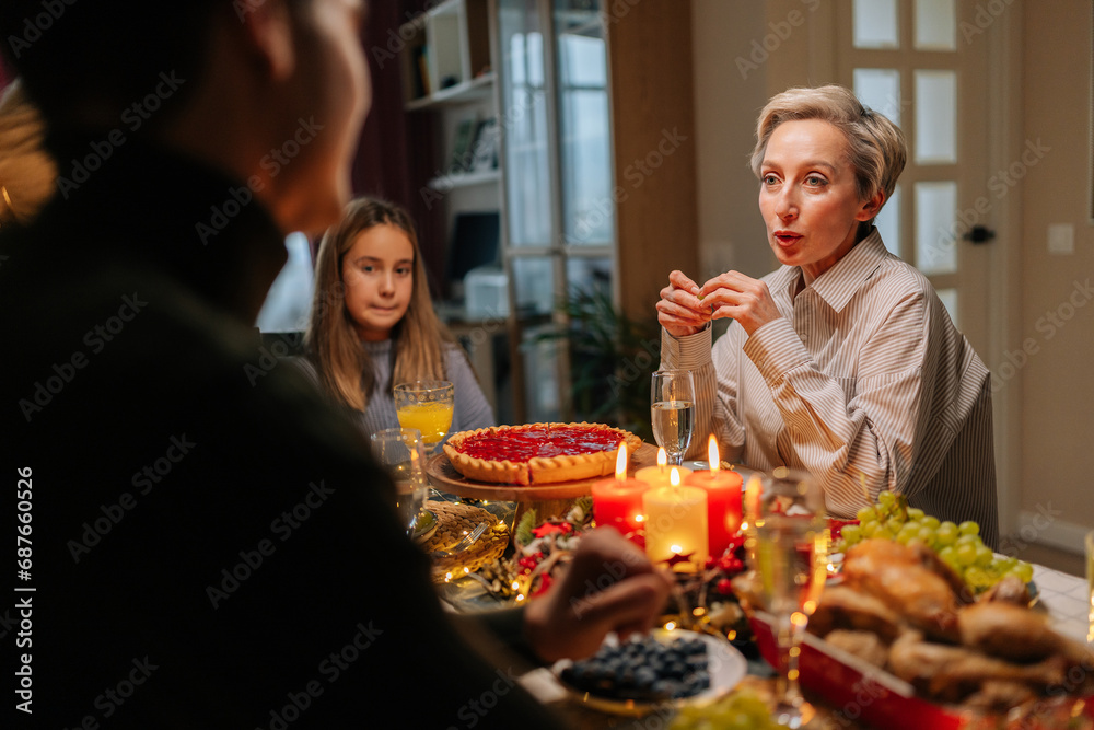 Elegant middle-aged mother-in-law celebrating Christmas with family, chatting at dinner holiday table in dark living room with cozy interior on xmas eve. Concept of home festive atmosphere.