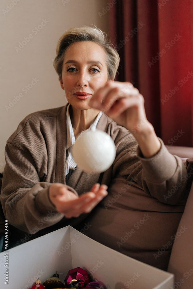 Vertical portrait of middle-aged woman holding Christmas ball, looking at camera with happy expression. Elegant female decorates xmas tree for New Year alone. Concept of home festive atmosphere. Vertical portrait of middle-aged woman holding Christmas ball, looking at camera with happy expression. Elegant female decorates xmas tree for New Year alone. Concept of home festive atmosphere.