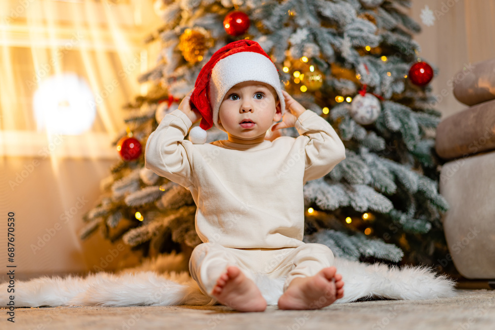 A Joyful Christmas Moment: A Child in a Santa Hat Admiring a Beautifully Decorated Christmas Tree. A small child wearing a santa hat sitting in front of a christmas tree A Joyful Christmas Moment: A Child in a Santa Hat Admiring a Beautifully Decorated Christmas Tree. A small child wearing a santa hat sitting in front of a christmas tree