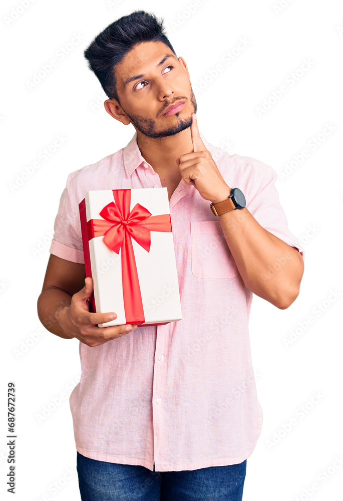 Handsome latin american young man holding gift serious face thinking about question with hand on chin, thoughtful about confusing idea Handsome latin american young man holding gift serious face thinking about question with hand on chin, thoughtful about confusing idea
