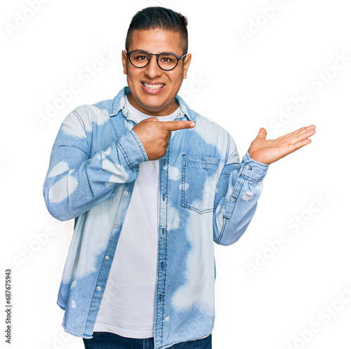 Young latin man wearing casual clothes and glasses amazed and smiling to the camera while presenting with hand and pointing with finger.