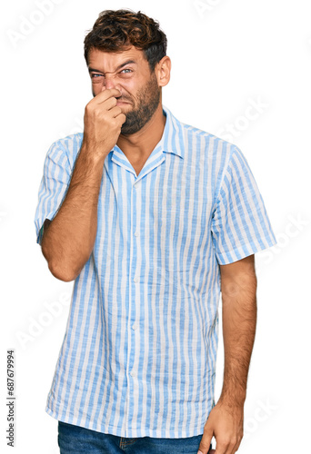 Handsome young man with beard wearing casual fresh shirt smelling something stinky and disgusting, intolerable smell, holding breath with fingers on nose. bad smell