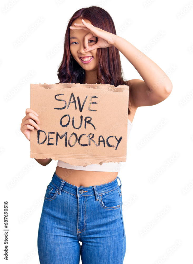 Young beautiful chinese girl holding save our democracy protest banner smiling happy doing ok sign with hand on eye looking through fingers Young beautiful chinese girl holding save our democracy protest banner smiling happy doing ok sign with hand on eye looking through fingers