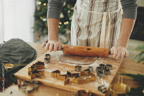 Hands kneading gingerbread dough with rolling pin, cooking spices, festive decorations on rustic table against stylish christmas tree. Man making christmas gingerbread cookies, holiday time