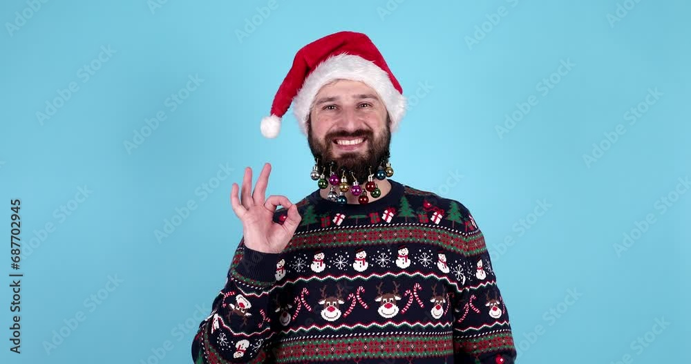 Happy excited bearded man in santa hat showing OK gesture and laughing, studio portrait over blue background. Christmas holidays concept. Ball toys in beard. Happy excited bearded man in santa hat showing OK gesture and laughing, studio portrait over blue background. Christmas holidays concept. Ball toys in beard.