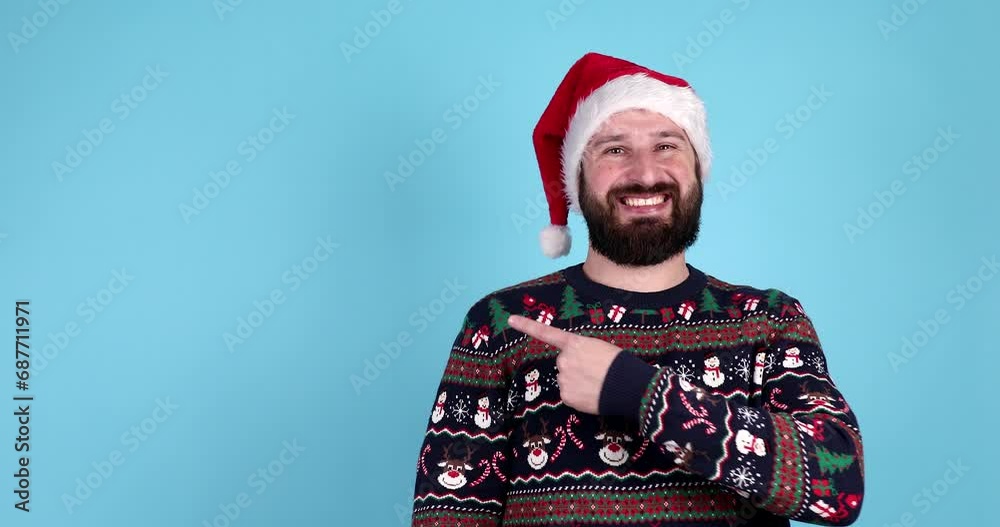 Young bearded male in Santa hat and warm christmas sweater is pointing up at something, winking and smiling while posing against blue background. Happy New Year, advertising. Close up, copy space. Young bearded male in Santa hat and warm christmas sweater is pointing up at something, winking and smiling while posing against blue background. Happy New Year, advertising. Close up, copy space.