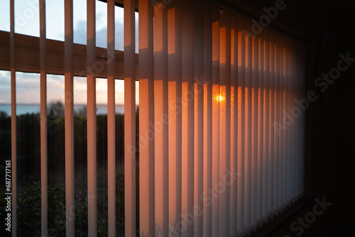 Shallow focus on vertical window blinds with a golden glow as the sun sets.There is a view of the sea and the glow of the setting sun.