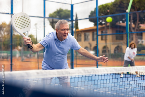 Focused aged man playing friendly paddleball match on outdoor summer court. Senior people sports concept..
