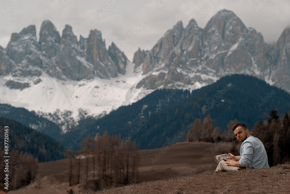 Fototapeta premium Young Attractive Man With Short-Cut Beard Taking A Well-Deserved Rest And Having A Delicious Meal After Exhausting Mountain Hike In Front Of Massive Alpine Rocks In The Dolomites Covered With Snow