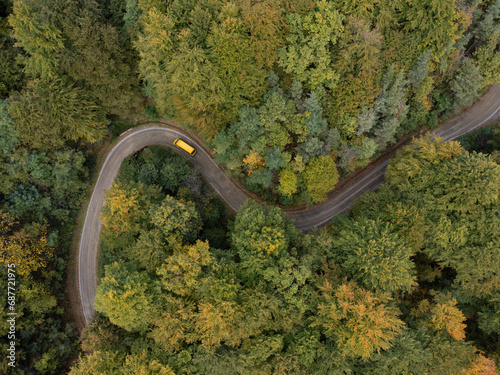 Bird view drone photo of curvy road through forest and yellow vehicle passing