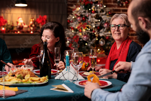 Grandmother at xmas dinner with people enjoying seasonal holiday at home with festive decorations and christmas tree. Old woman meeting with relatives and friends around the table.