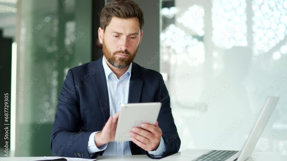 Businessman in formal suit is using digital tablet while sitting at workplace in business office. Worker reads or typing message, chats with a client, banking, checks email or swiping browsing online Businessman in formal suit is using digital tablet while sitting at workplace in business office. Worker reads or typing message, chats with a client, banking, checks email or swiping browsing online