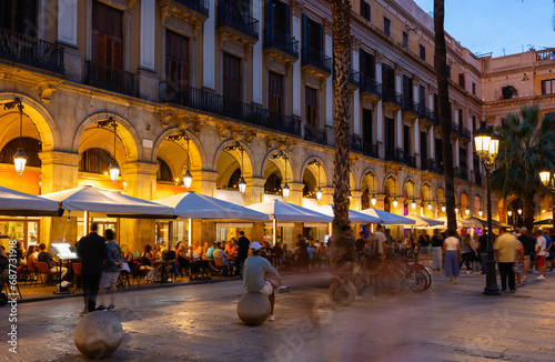 Evening view of bustling Placa Reial in Barcelona with well-lit cafes and people enjoying leisurely stroll. Favorite place for locals and tourists alike