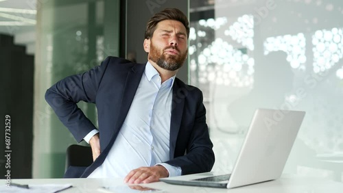 Tired businessman in formal suit suffering from back pain while working on laptop sitting at workplace in business office. Upset man massages painful lower back muscles He is exhausted and overworked photo