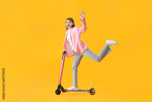 Portrait of excited businesswoman riding kick scooter and waving hand on yellow background © Pixel-Shot