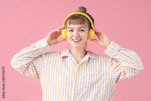 Portrait of pretty young woman in pajamas and headphones listening to music on pink background