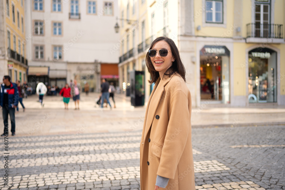 Fototapeta premium Smiling woman standing crosswalk on street and posing with sunglasses. Woman in coat and sunglasses standing sideways at intersection with blur background. Female looking at camera and smiling