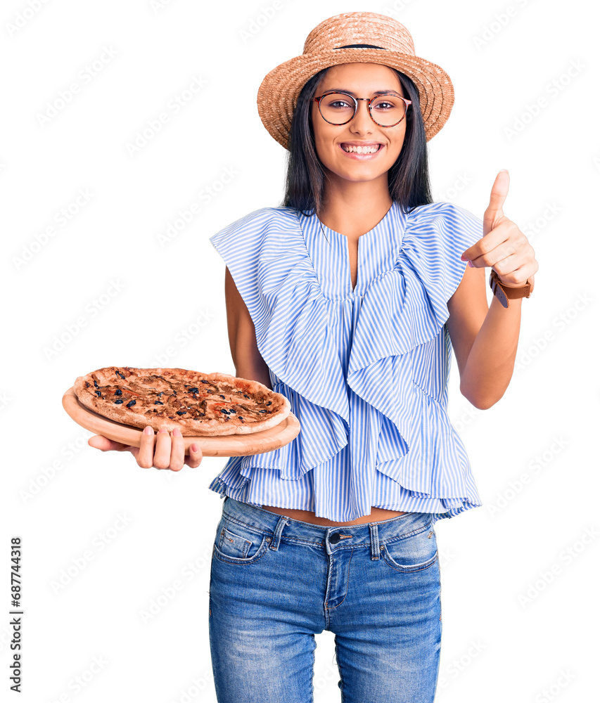 Young beautiful latin girl wearing summer hat and glasses holding pizza smiling happy and positive, thumb up doing excellent and approval sign Young beautiful latin girl wearing summer hat and glasses holding pizza smiling happy and positive, thumb up doing excellent and approval sign