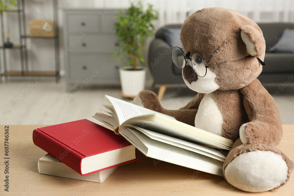 Teddy bear with glasses and open book on wooden table in room, closeup - obrazy, fototapety, plakaty 