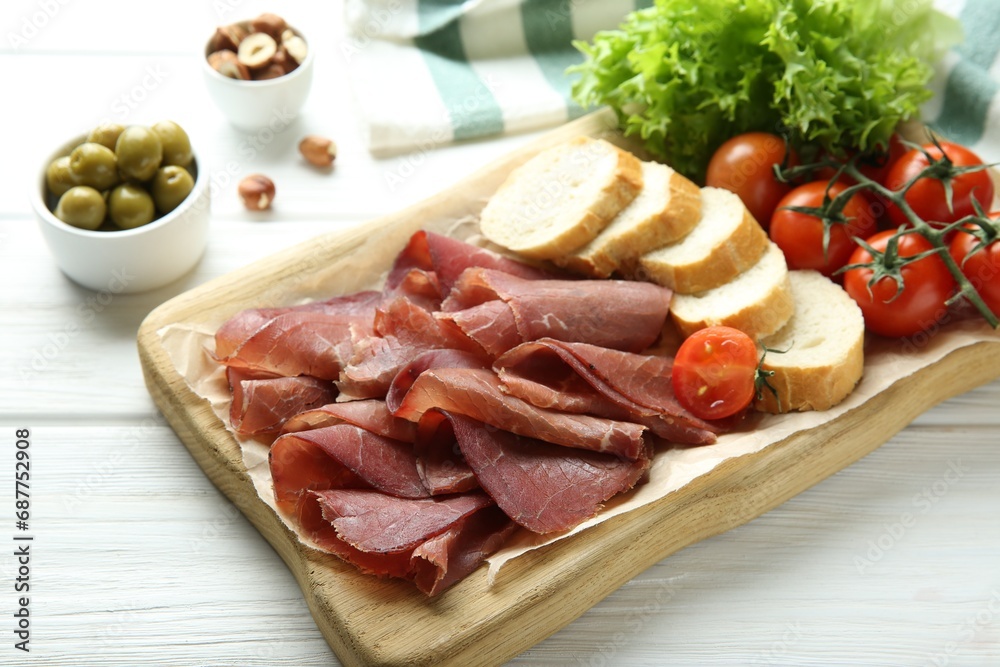 Board with delicious bresaola served with bread and tomato on white wooden table Board with delicious bresaola served with bread and tomato on white wooden table