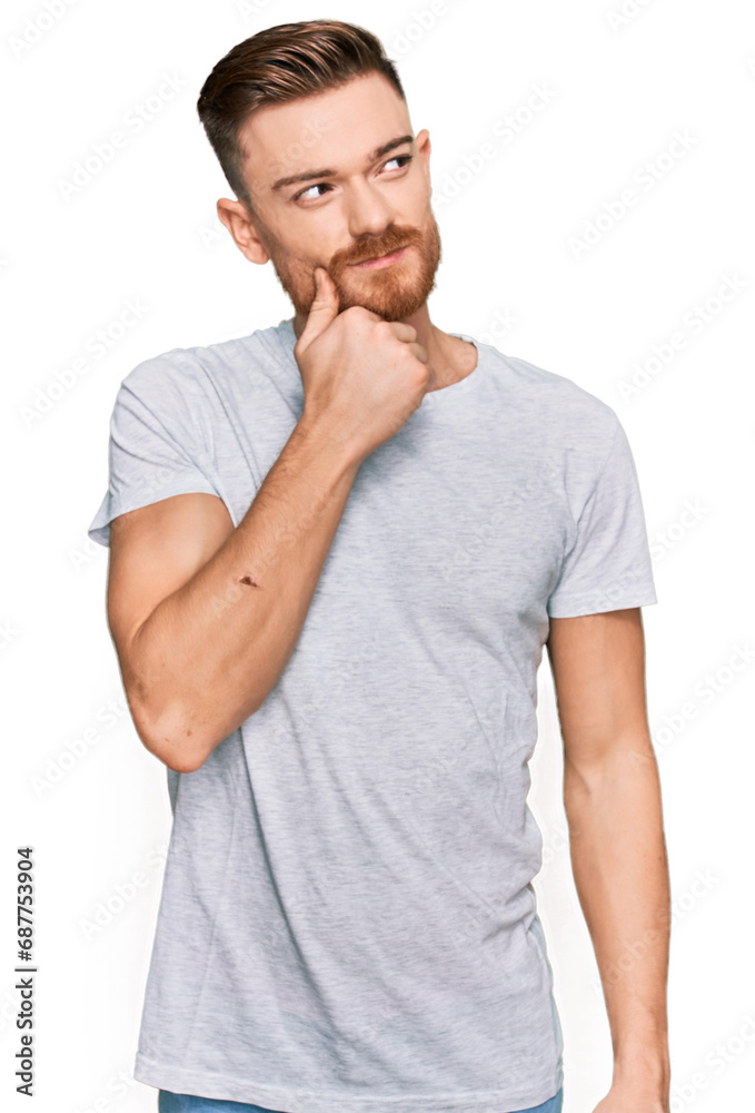 Young redhead man wearing casual grey t shirt with hand on chin thinking about question, pensive expression. smiling with thoughtful face. doubt concept. Young redhead man wearing casual grey t shirt with hand on chin thinking about question, pensive expression. smiling with thoughtful face. doubt concept.