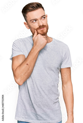 Young redhead man wearing casual grey t shirt with hand on chin thinking about question, pensive expression. smiling with thoughtful face. doubt concept.